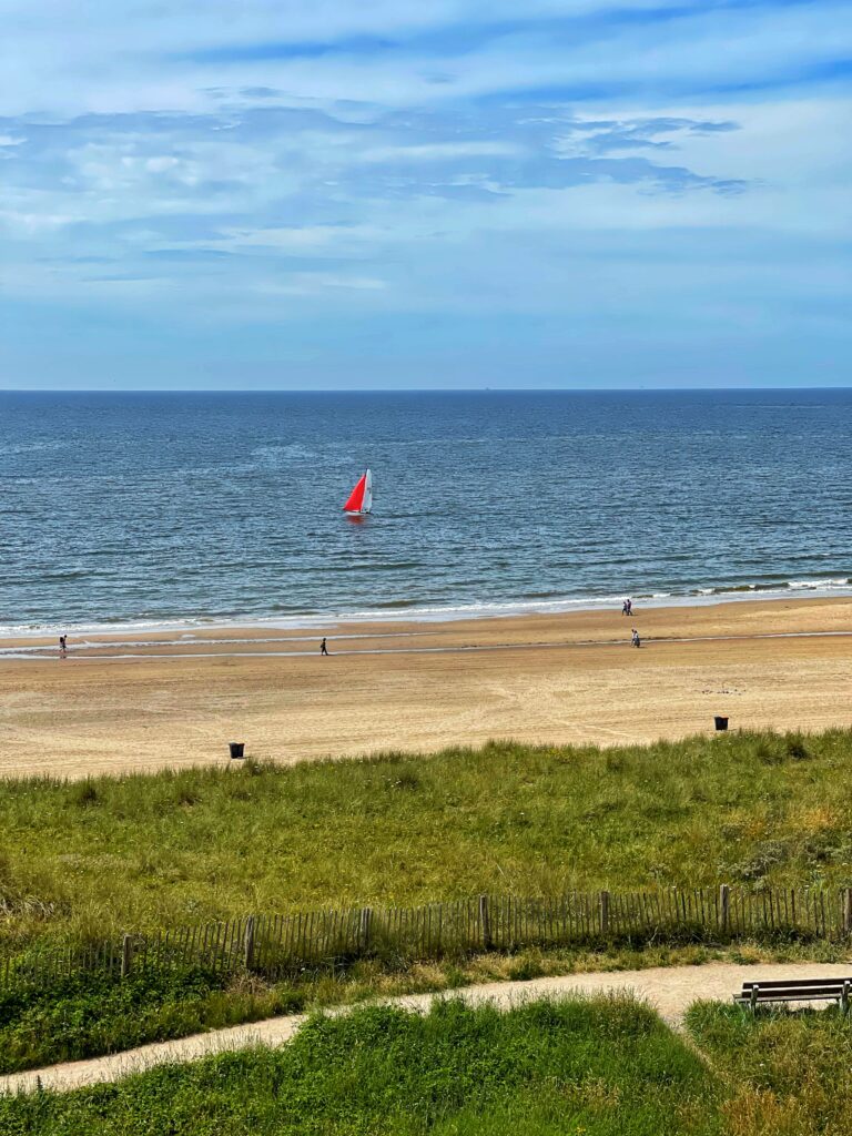 Dünen, dahinter Strand mit einzelnen Spaziergängern, das Meer mit einem Segelboot mit einem orangefarbenen und einem weißen Segel und blauer Himmel mit ein paar Wolken.