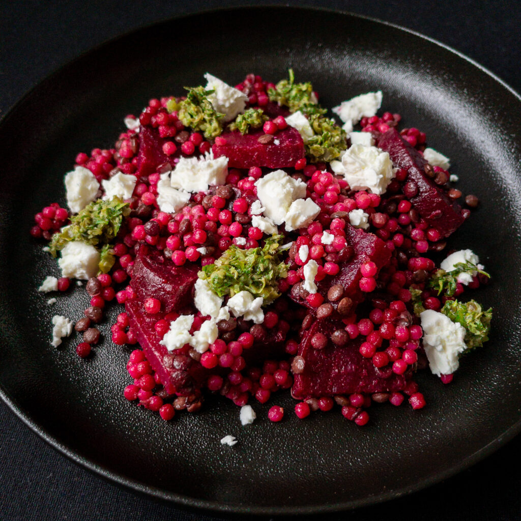 Ein schwarzer Teller auf schwarzem Untergrund. Darauf eine Portion Rote Bete-Salat mit vom Saft rosa gefärbten kleinen Nudelkügelchen (Fregola Sarda) und braunen Linsen, getoppt von weißen Feta-Bröckchen und Klecksen mit grüner Salsa Verde.