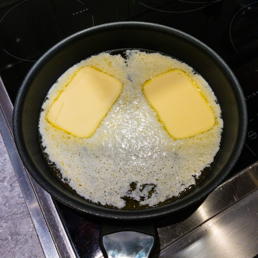 Blick von oben in ein Pfanne auf dem Herd, in der 2 Stücke Butter schmelzen.