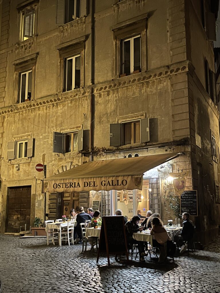 Nachts in Rom. Unter einer Markise einer Osteria sitzen Menschen und essen. Alles ist warm beleuchtet und sieht so gemütlich aus.