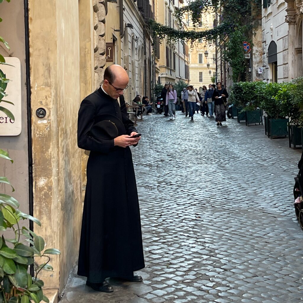 Ein Priester im Talar steht am Rande einer Altstadtgasse und tippt in sein Handy. Zwei Welten prallen aufeinander…