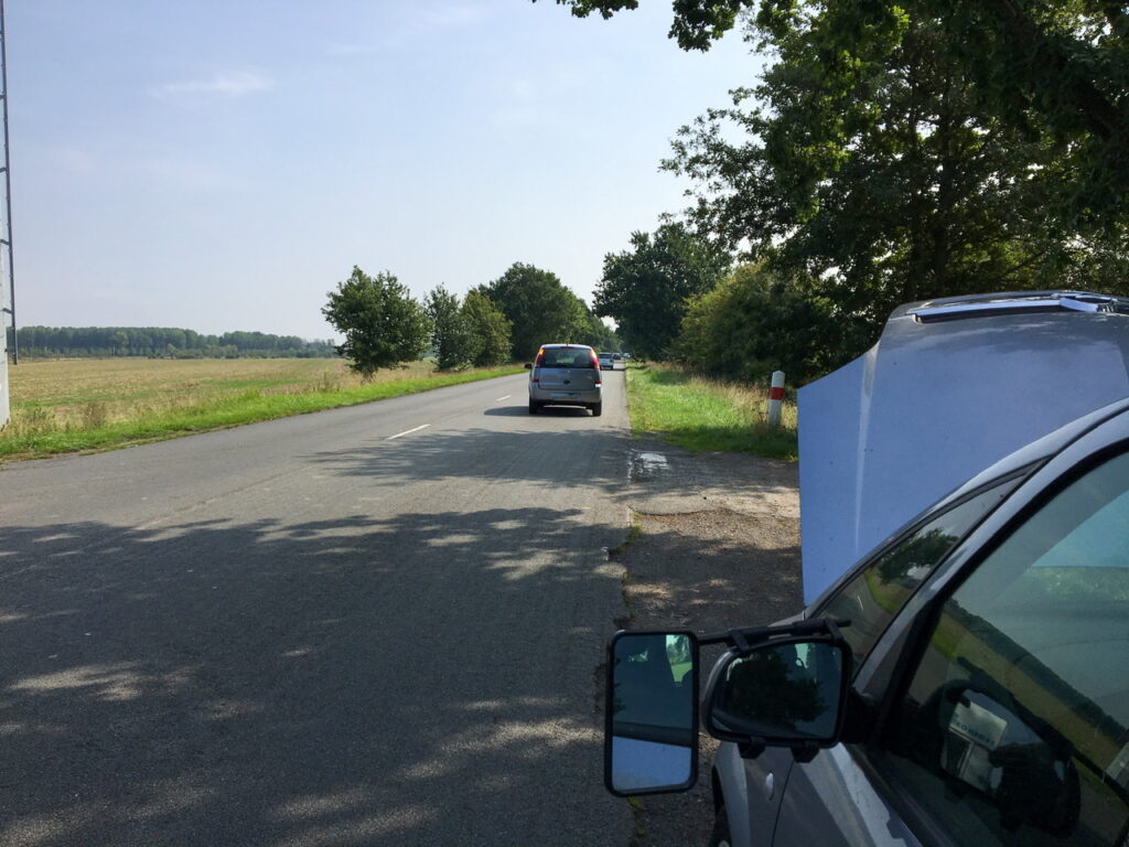 Unser kaputtes Auto steht mit offener Motorhaube am Straßenrand einer französischen Landstraße. Es ist Sommer und die Sonne schein.
