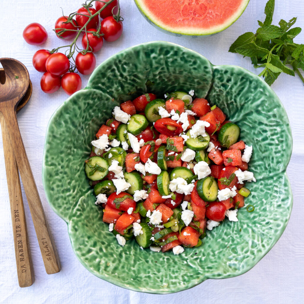 Auf einer weißen Tischdecke steht eine grüne Keramik-Salatschüssel. Darin der Wassermelonensalat mit Tomaten und Gurken, Kräutern und Feta. Links daneben eine Salatbesteck aus Holz und im Uhrzeigersinn angerichtete Deko aus Strauchtomaten, ein Stück Wassermelone und Minze.