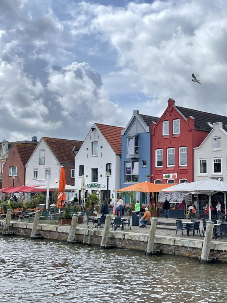 Die Uferpromenade in Husum. Kleine bunte Häuser in Reihe, davor Außengastronomie und das Hafenbecken. Wolkenhimmel und eine Möwe fliegt darüber.