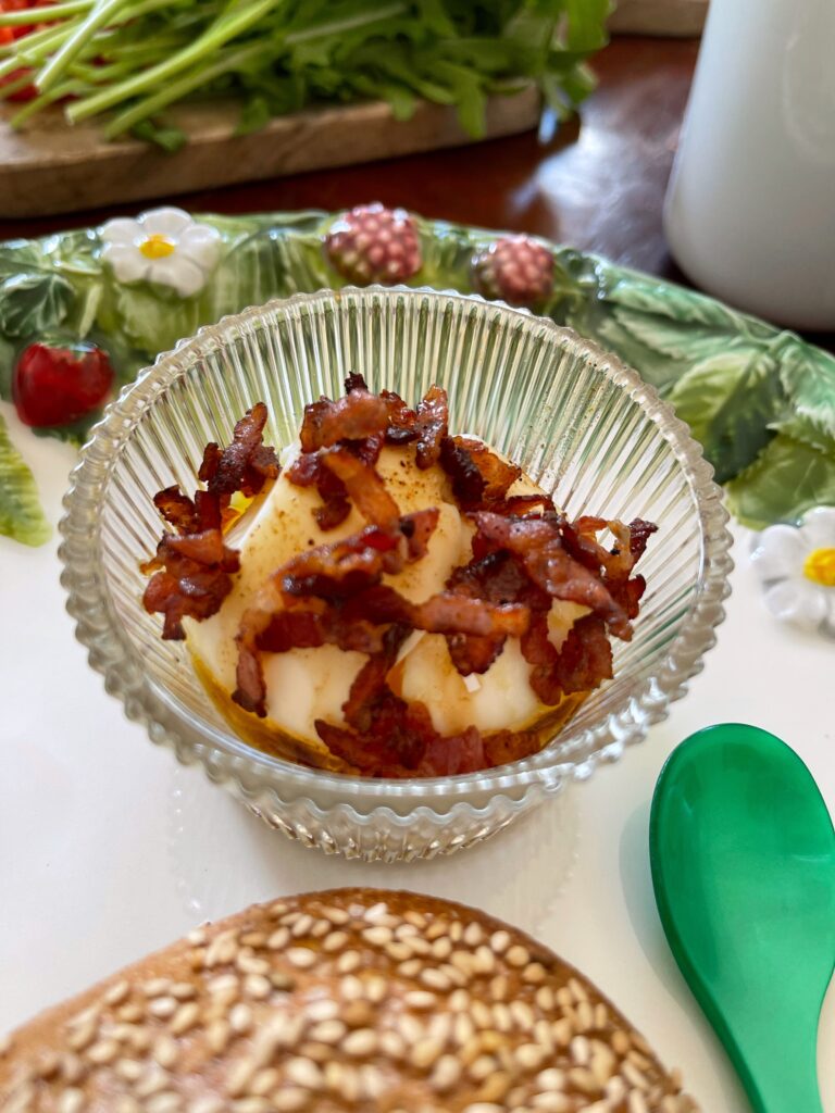 Blick auf einen Frühstücksteller. Vorne liegt ein Sesambrötchen, dahinter steht ein kleines, geriffeltes Glas mit einem weichgekochten Ei, bedeckt mit kross gebratenen Speckwürfeln, darin. Daneben liegt ein Eierlöffel. Im Hintergrund steht ein Brett mit Rucola und roter Paprika und eine Kaffeetasse.