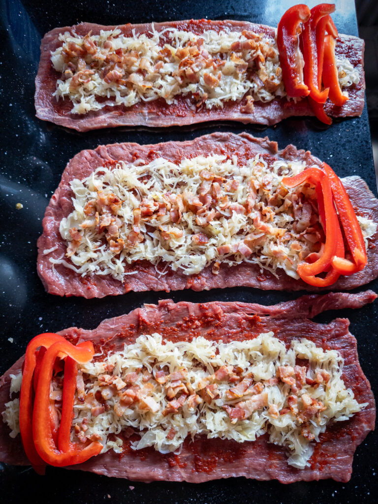 Blick auf die vorbereiteten Rouladen. Man sieht die Sauerkraut-Speck-Füllung und oben quer die roten Paprikastreifen.