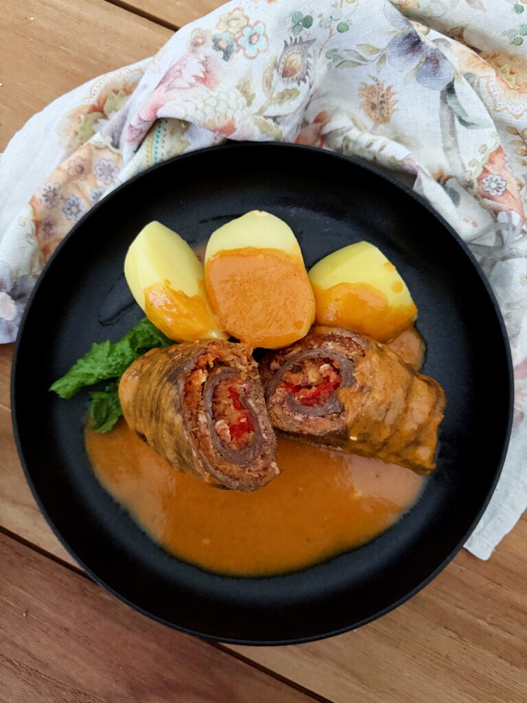 Blick auf einen schwarzen Teller, darauf eine in der Mitte quer durchgeschnittene Rindfleischroulade gefüllt mit Sauerkraut und Paprika auf einem Saucenspiegel mit Salzkartoffeln und etwas dekorativem Grün.