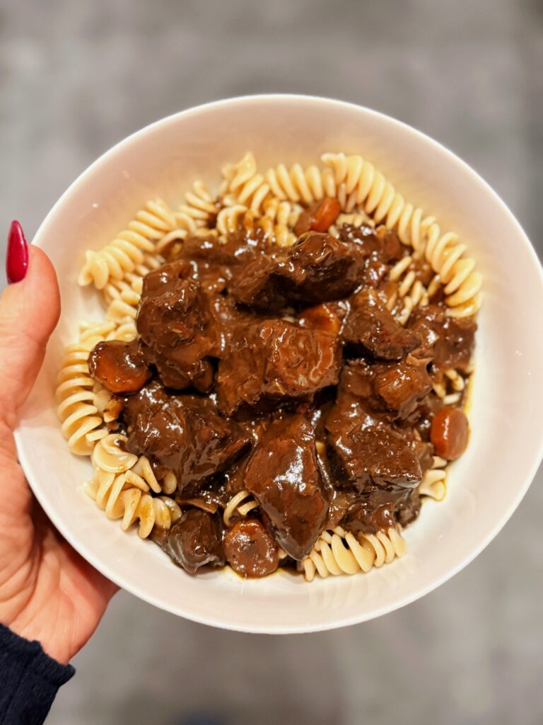 Eine Hand hält einen Teller auf dem sich gekochte Spiralnudeln mit Wildgulasch in dunkel glänzender Sauce befindet.